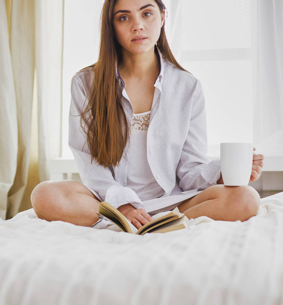 Woman sitting in bed reading a book and having breakfast. - Foto, Imagen