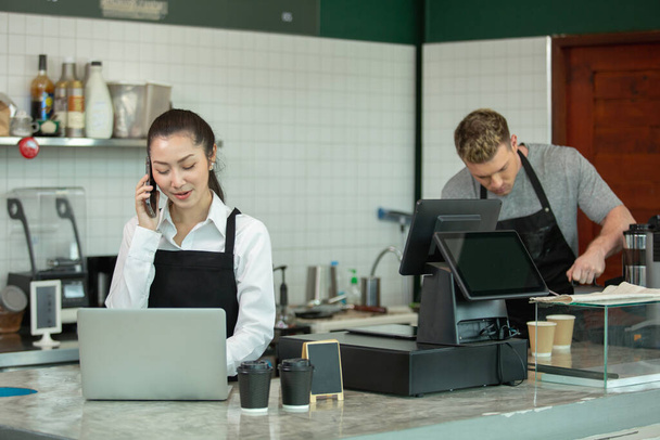 Asijské mladé ženy barista nosit zástěru mluvení telefon a používání notebooku v kavárně. ženský personál stojící u pultu přijímá objednávky zákazníků v kavárně. obchodní kavárna koncept. - Fotografie, Obrázek
