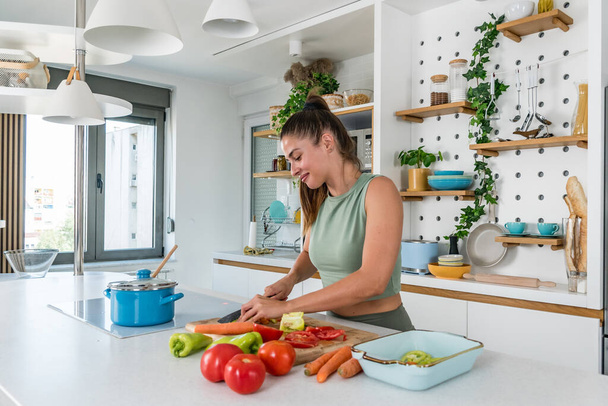 Young sporty woman cutting and cooking fresh natural vegetables at kitchen and cooking for healthy lunch. Self care yoga nutritionist girl having healthy meal after workout. - Valokuva, kuva