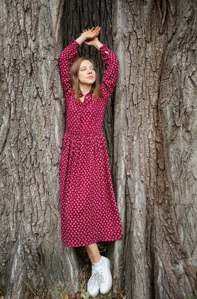 Full length portrait of a girl in a long burgundy dress posing in the shade of an oak tree trunk. Loneliness, contemplation, solitude, fantasies alone - Fotografie, Obrázek