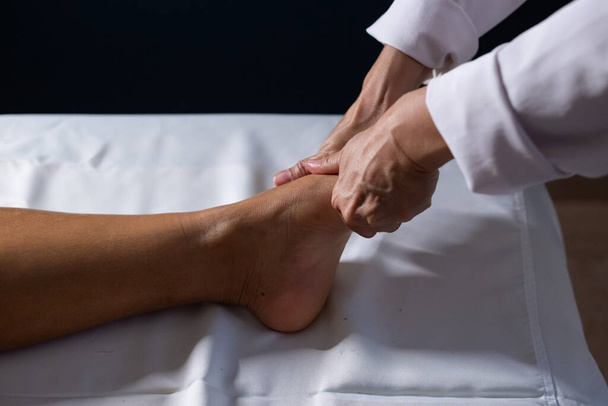 Detail of masseuse hands applying therapeutic massage on the foot of a patient who is lying on a stretcher with white sheet. - Foto, Imagem