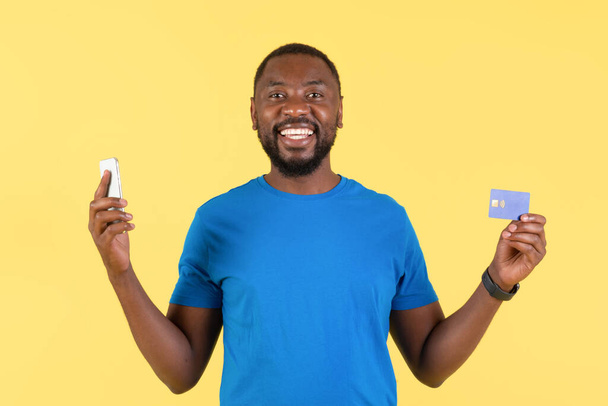 Усміхнений афроамериканський чоловік тримає мобільний телефон і кредитні картки покупки онлайн в додатку, що стоїть в студії над жовтим тлом. Мобільний банкінг додаток та концепція електронної комерції
 - Фото, зображення