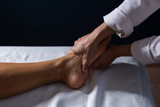 Деталь рук массажистки нанесения лечебного массажа на ногу пациента, лежащего на носилках с белым полотном. - Фото, изображение