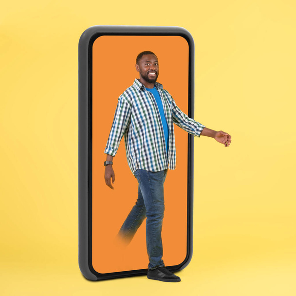 Cheerful Black Guy Stepping Out Of Huge Smartphone Screen Walking Over Yellow Studio Background. Man Advertising Great Mobile Offer And Application. Full Length, Square - Fotoğraf, Görsel