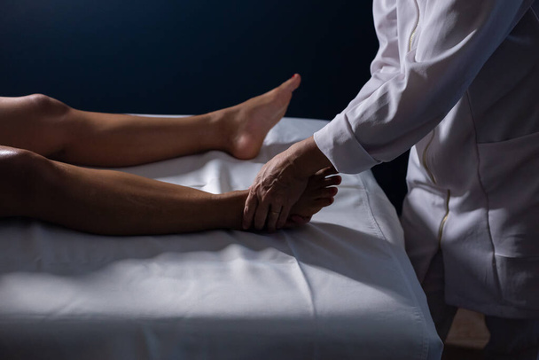 Деталь рук массажистки нанесения лечебного массажа на ногу пациента, лежащего на носилках с белым полотном. - Фото, изображение