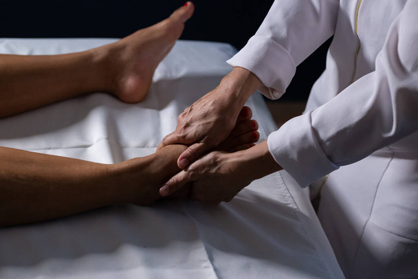 Detail of masseuse hands applying therapeutic massage on the foot of a patient who is lying on a stretcher with white sheet. - Fotó, kép