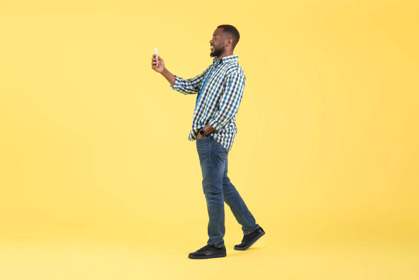 African American Male Using Phone Posing Walking On Yellow Background In Studio. Full Length Shot Of Millennial Man Holding Cellphone Using Mobile Application And Texting. Side View - Foto, immagini
