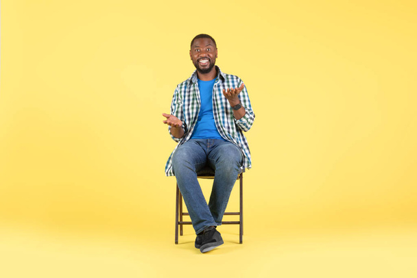 Happy African American Male Talking Looking At Camera Sitting In Chair Over Yellow Background. Guy Communicating Posing In Studio. Full Length Shot. Modern Communication Concept - Foto, Bild