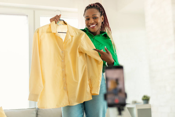 Happy african american female fashion blogger showing yellow shirt and looking at smartphone camera, recording review of new collection - Photo, image