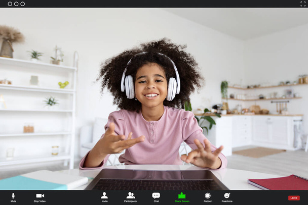 Screenshot of cheerful african american school girl with bushy hair having online class, using laptop and wireless headphones, smiling and gesturing at camera, home background. Online education - Foto, Bild