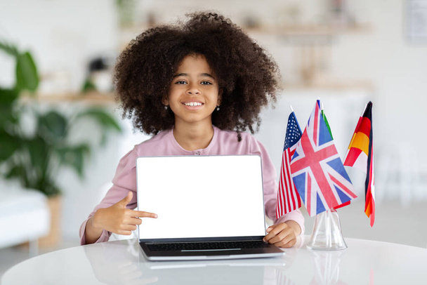Happy black school girl with bushy hair schooler sitting at desk with international flags, showing computer with white empty screen, child learning foreign languages online, mockup, copy space - Foto, immagini