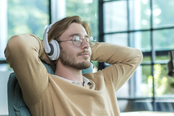 A man in an office on a chair with his arms folded is listening to music in headphones resting, a blond businessman is working inside an office building - Foto, Imagem