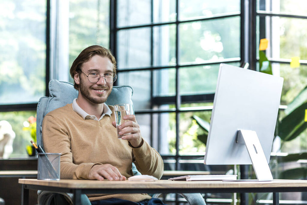 Young successful businessman man in modern office at work smiling and looking at camera, businessman holding glass of water, worker in glasses and casual clothes - Foto, Imagen