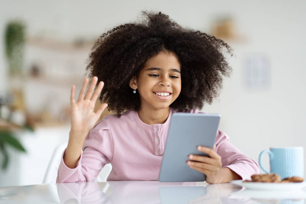 Cheerful african american girl with bushy hair having video call, sitting at desk, eating cookies, drinking juice or cocoa, holding digital tablet, waving at pad screen and smiling, copy space - Photo, image