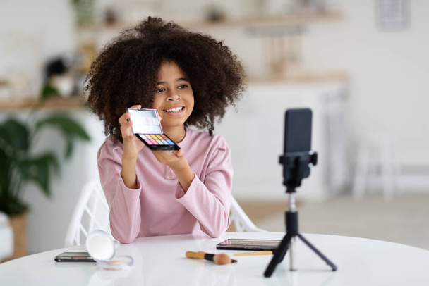 Beauty blog for kids concept. Cheerful cute black preteen girl doing makeup and recording vlog on phone set on tripod, child sitting at table at home, showing fancy eyeshadows for little girls - Fotó, kép