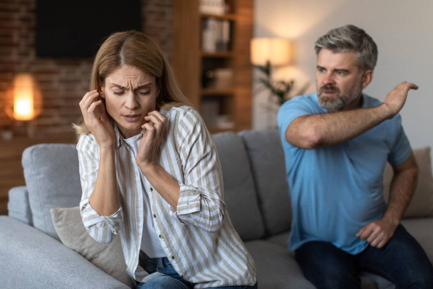 Aggressive nervous middle aged caucasian man with beard scolds and wants to hit sad scared wife, fighting in living room interior. Relationship problems, stress at home, abuse and domestic violence - Photo, Image