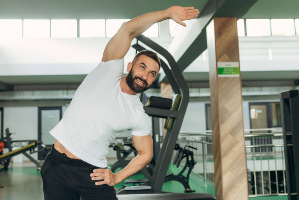 Portrait of an athlete in the gym. A man wearing a white T-shirt is smiling and doing exercises - Photo, Image