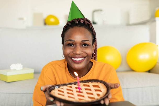 З днем народження тебе. Щаслива афроамериканка у святковому капелюсі, що тримає пиріг з палаючою свічкою і посміхається до камери, сидячи вдома. Святкування свят - Фото, зображення