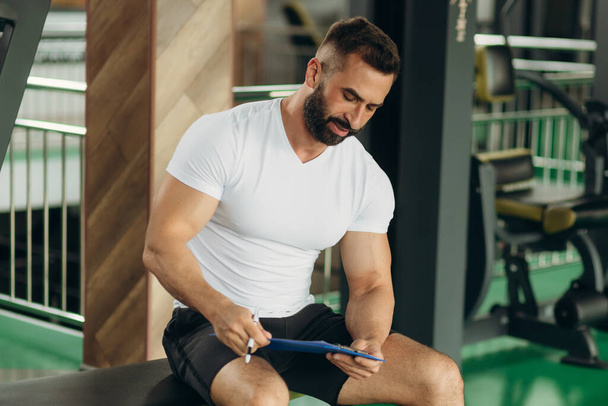 A young fitness instructor is reading a training plan in the gym. Dressed in a white T-shirt. - Foto, imagen