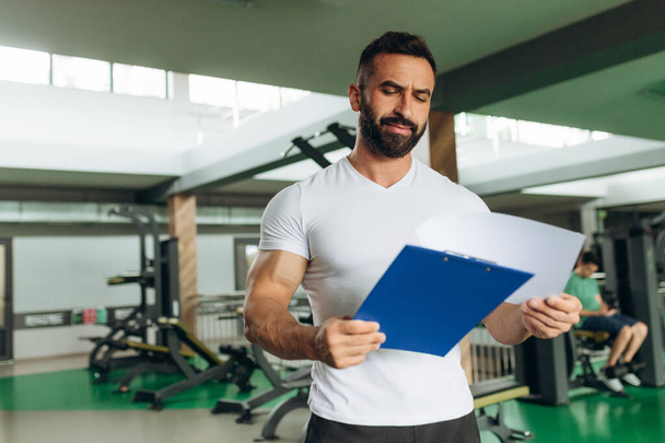 Entrenador musculoso guapo que mira el plan de acondicionamiento físico en el portapapeles para hacer ejercicio en el gimnasio
 - Foto, Imagen