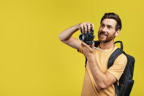 Positive middle aged man with backpack taking picture on professional DSLR camera over yellow studio background, free space. Male tourist shooting travel photos during his adventures - Φωτογραφία, εικόνα