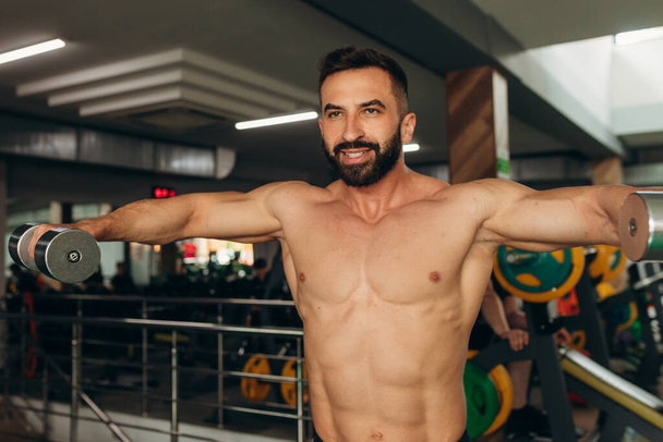 Αθλητικός άνθρωπος κάνει ασκήσεις με αλτήρες με γυμνό κορμό στο γυμναστήριο - Φωτογραφία, εικόνα