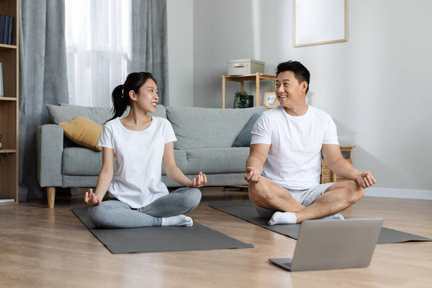 Счастливые муж и жена азиатской семьи в спортивной одежде сидя на фитнес-коврики в гостиной на дому, имея утреннюю йогу вместе, с помощью ноутбука, посещая онлайн курс йоги, копировать пространство - Фото, изображение