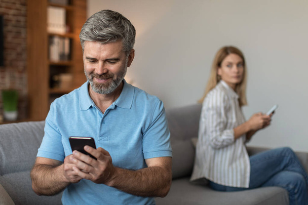 Happy adult caucasian husband with beard plays on smartphone, suffers from gadget addiction, ignores wife in living room interior. Distrust, relationship problems, emotions, cheating and gambling - Photo, image