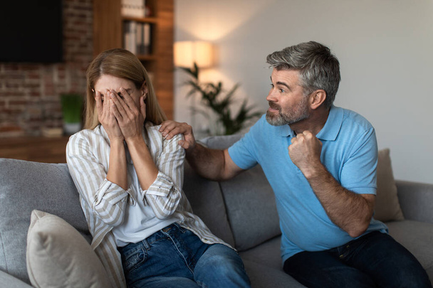 Angry aggressive mature caucasian husband with beard threatens to scared wife with fists in living room interior. Problems in relationships, emotions, quarrel at home, beating and domestic violence - Foto, imagen