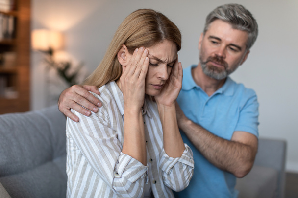 Sad adult caucasian man with beard calm offended wife, lady suffers from migraine in living room interior. Pain and headache, bad news, menopause, relationships problems, depression and stress at home - Photo, Image