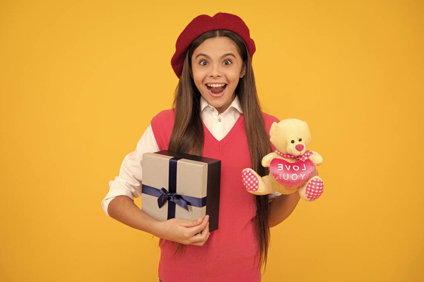 Fransız bereli hayretler içinde genç bir kız oyuncağı elinde tutuyor ve sarı arka planda hediye kutusu, hayret verici.. - Fotoğraf, Görsel