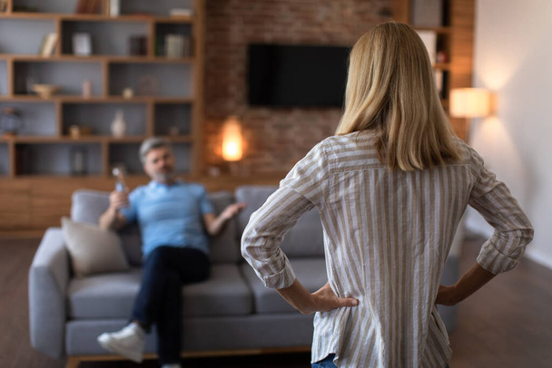Unhappy sad adult caucasian wife angry at drunk husband with bottle in living room interior, back, blurred. Relationship problems, alcoholism, quarrel and addiction at home, conflict due covid-19 - Fotografie, Obrázek