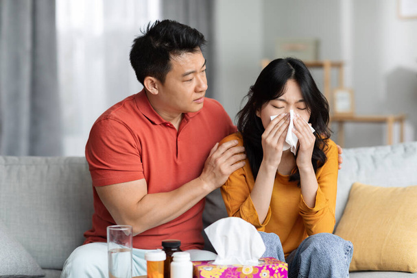 Loving asian husband taking care of sick wife sitting on sofa next to tea table full of medicine and sneezing nose, loving man hugging his ill girlfriend. Cold, flu, coronavirus concept - Fotoğraf, Görsel