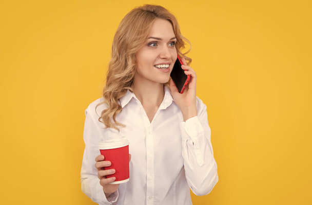 heureuse femme blonde avec tasse de café parlant au téléphone sur fond jaune. - Photo, image