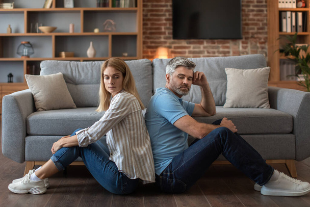 Unhappy mature caucasian male with beard ignores offended woman, sit on floor back to back in living room interior. Quarrel, relationship problems, people emotions, depression and conflict at home - 写真・画像