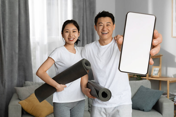 Sporty asian husband and wife in sportswear holding fitness mats in living room, couple having morning yoga at home, showing smartphone with white blank screen, using yoga app, copy space, mockup - Fotó, kép