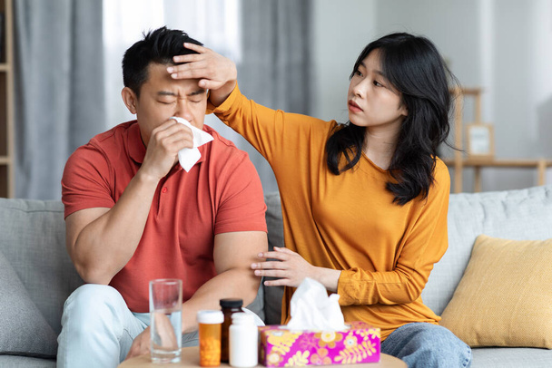 Young asian wife taking care of sick husband sitting on sofa next to tea table full of medicine and sneezing nose, loving woman touching her ill boyfriend forehead. Cold, flu, coronavirus concept - Photo, image
