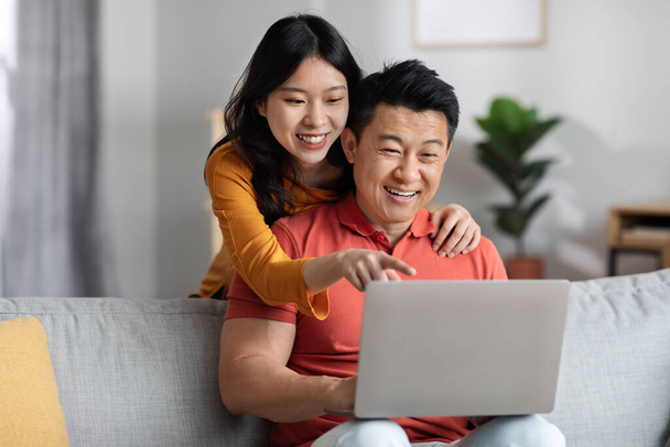 Loving chinese spouses shopping on Internet from home, cheerful young asian woman hugging her husband from behind, pointing at laptop screen, family choosing new furniture online, copy space - Foto, Imagen