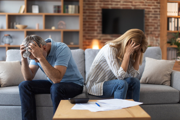 Unhappy mature caucasian couple holding heads with hands, suffer from debts in room interior with documents and calculator. Taxes, problems with bills, home bookkeeping, credit, bankruptcy and stress - 写真・画像