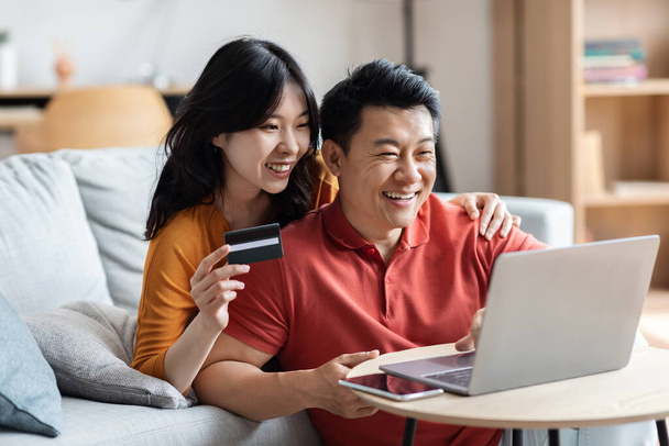 Cheerful chinese man and woman embracing, using modern laptop and plastic credit card, happy asian spouses paying for goods and services online while staying home, copy space - Photo, Image