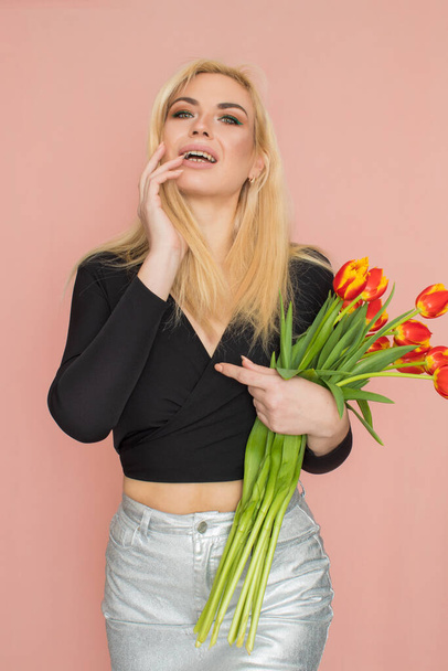 Fashion model woman in fashionable clothes on pink background. Wearing stylish clothing, black blouse, silver skirt. Posing in studio. Holding red tulips in her hands - Foto, Imagen