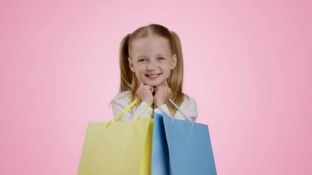 Joy of sales. Studio portrait of cute little girl shopaholic enjoying success, carrying blue and yellow shopping bags, pink background, slow motion - Felvétel, videó
