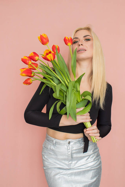 Fashion model woman in fashionable clothes on pink background. Wearing stylish clothing, black blouse, silver skirt. Posing in studio. Holding red tulips in her hands - Photo, image