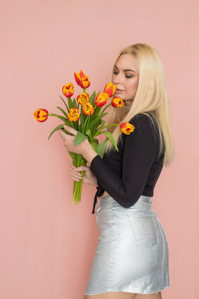Fashion model woman in fashionable clothes on pink background. Wearing stylish clothing, black blouse, silver skirt. Posing in studio. Holding red tulips in her hands - Φωτογραφία, εικόνα