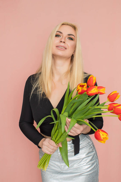 Fashion model woman in fashionable clothes on pink background. Wearing stylish clothing, black blouse, silver skirt. Posing in studio. Holding red tulips in her hands - Foto, imagen