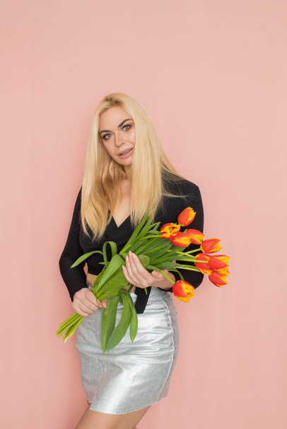 Fashion model woman in fashionable clothes on pink background. Wearing stylish clothing, black blouse, silver skirt. Posing in studio. Holding red tulips in her hands - Фото, зображення