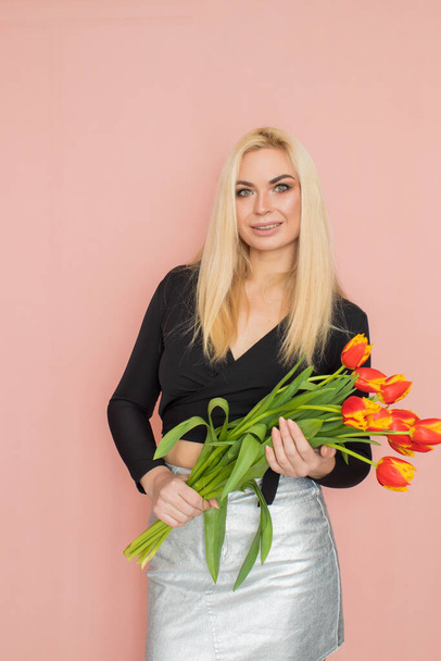 Fashion model woman in fashionable clothes on pink background. Wearing stylish clothing, black blouse, silver skirt. Posing in studio. Holding red tulips in her hands - Foto, imagen