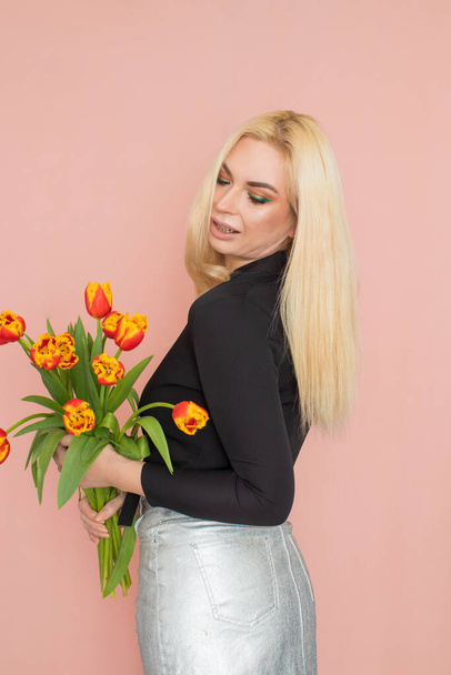 Fashion model woman in fashionable clothes on pink background. Wearing stylish clothing, black blouse, silver skirt. Posing in studio. Holding red tulips in her hands - Фото, изображение