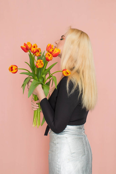 Fashion model woman in fashionable clothes on pink background. Wearing stylish clothing, black blouse, silver skirt. Posing in studio. Holding red tulips in her hands - Photo, Image