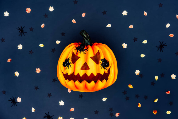 Top view bright Halloween background with carved orange pumpkin. Halloween Jack-o-lantern with spiders on it and multicolored autumn tinsel on the dark blue background. Flat lay. Selective focus - Φωτογραφία, εικόνα